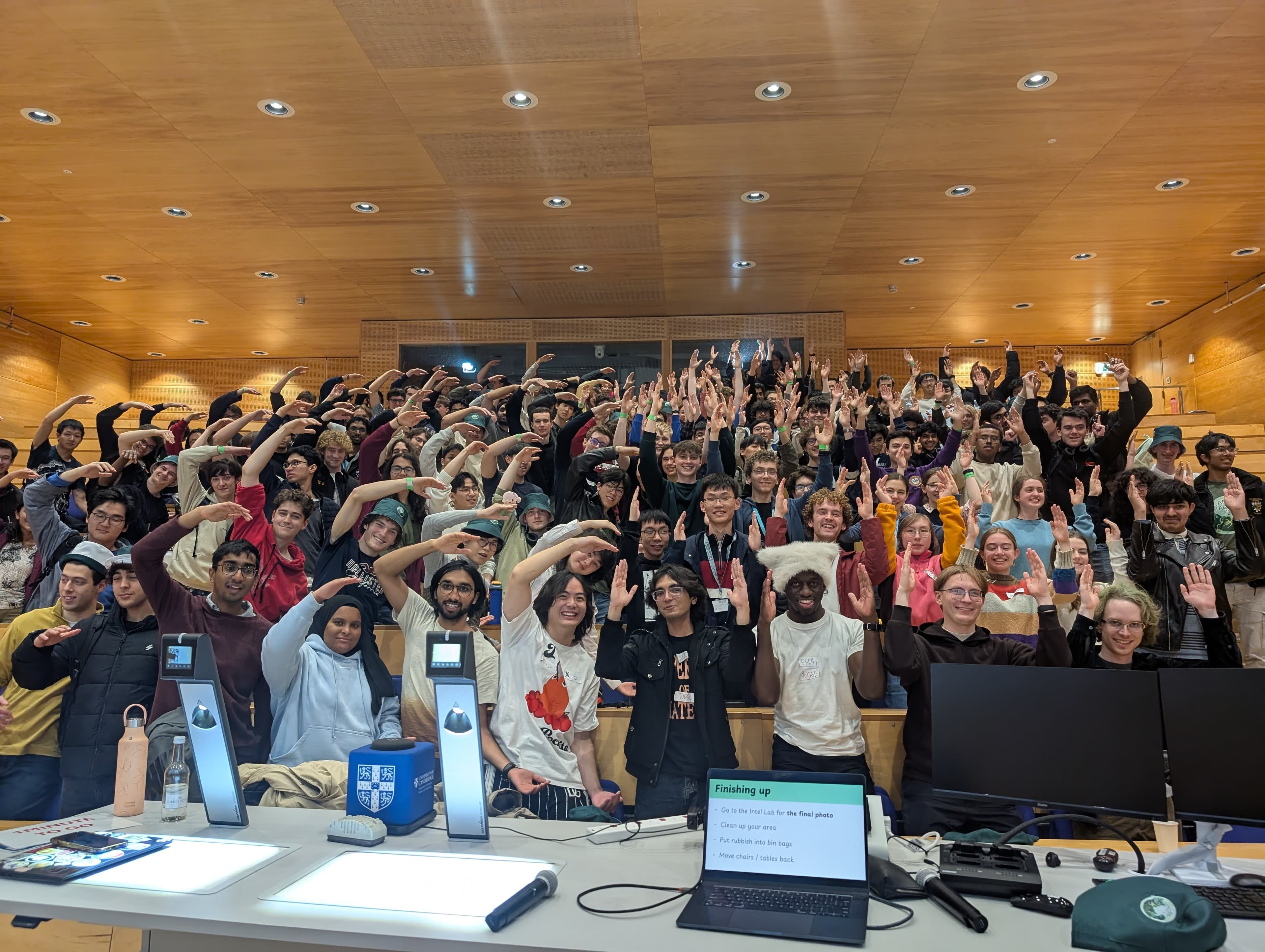 Group photo of all CamHack 2025 participants packed into the lecture theatre with arms raised